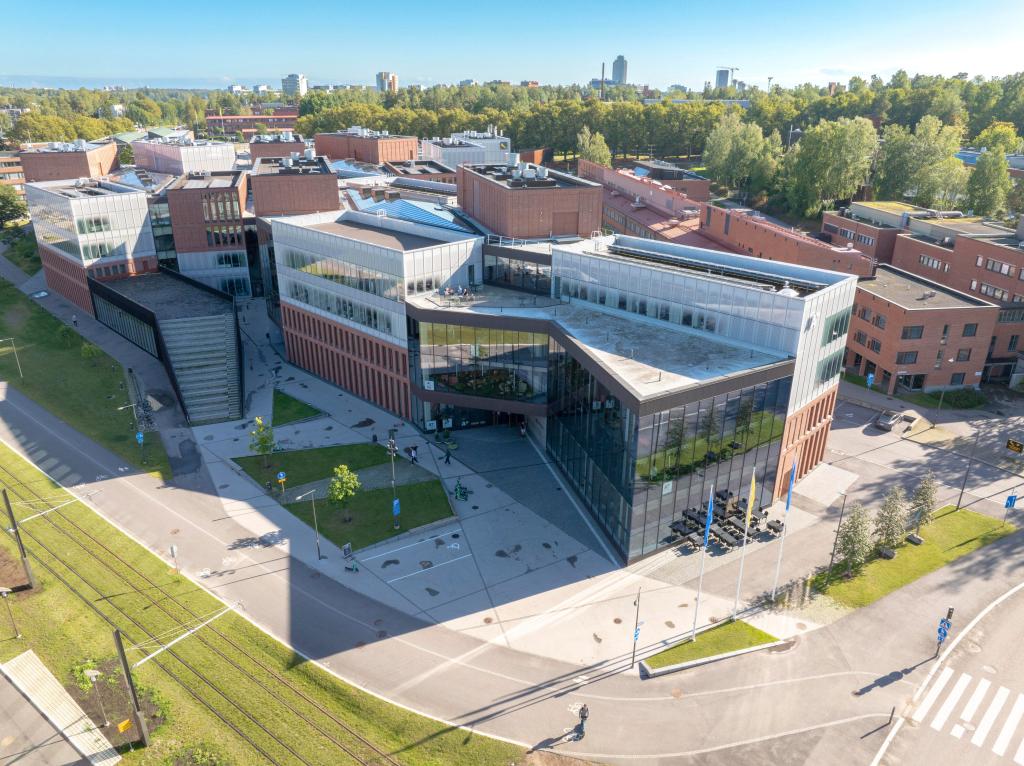 Aerial view of the Aalto University School of Business, a building of red brick and glass with flags at its main entrance, on a sunny summer day.