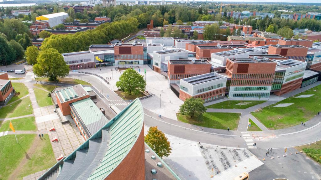Aerial view of the Otaniemi campus: the copper roof of Alvar Aalto's main building in the foreground, the red-brick Väre building and its square behind it, surrounded by trees.